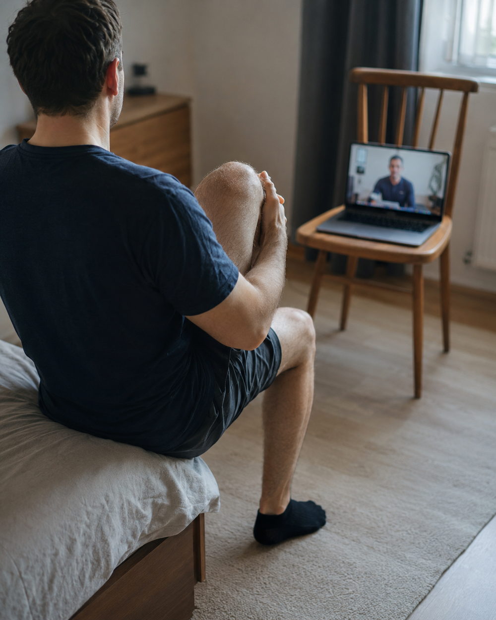 A patient seated at home doing a guided knee movement while a physiotherapist watches on a laptop video call